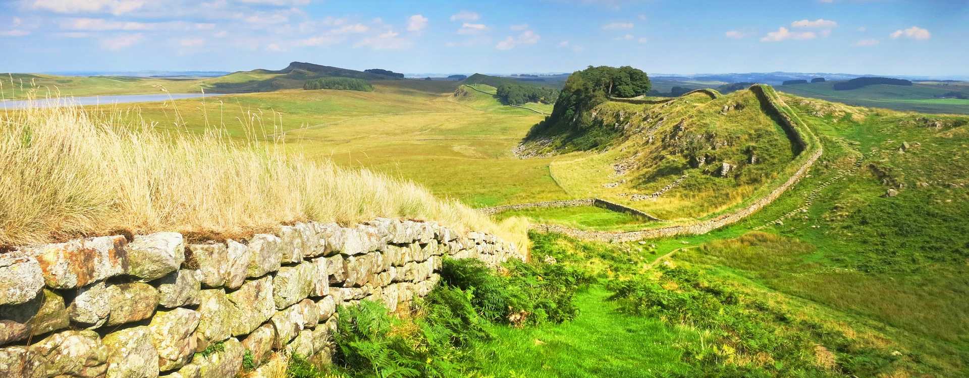 Wide panoramic view of the preserved stone ruins of Hadrian's Wall curving along the high ridges of the Whin Sill escarpment in the British countryside – Travel guide for Roman wall ticket bookings in 2026