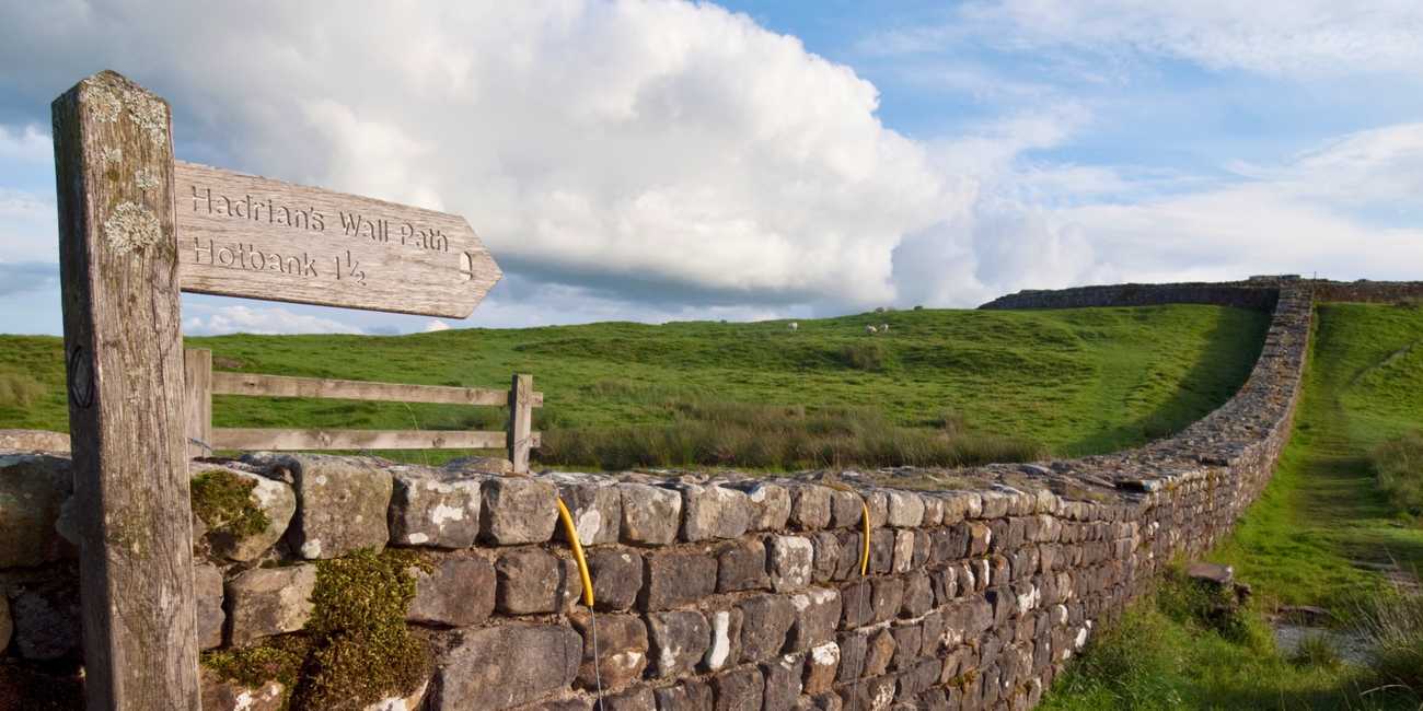 A rustic wooden fingerpost sign pointing toward the Hadrian's Wall Path and Hotbank mounted beside a historic dry stone wall running up a green pasture hill