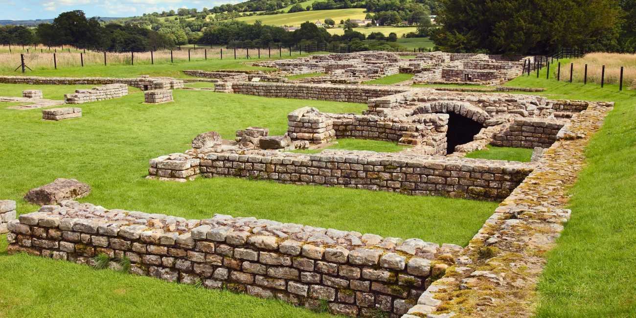 Excavated stone walls of the central headquarters building and vaulted underground strongroom stairs at Chesters Roman Fort under a cloudy sky.